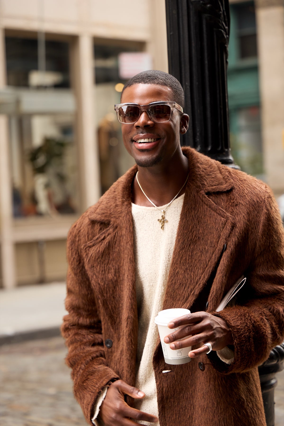 Man in a brown shearling coat and sunglasses holding a coffee